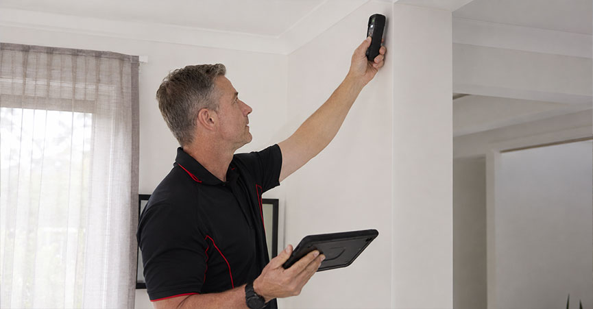 A building inspector holding a digital tablet while using a moisture meter device near the ceiling to check for leaks and dampness during a home inspection.